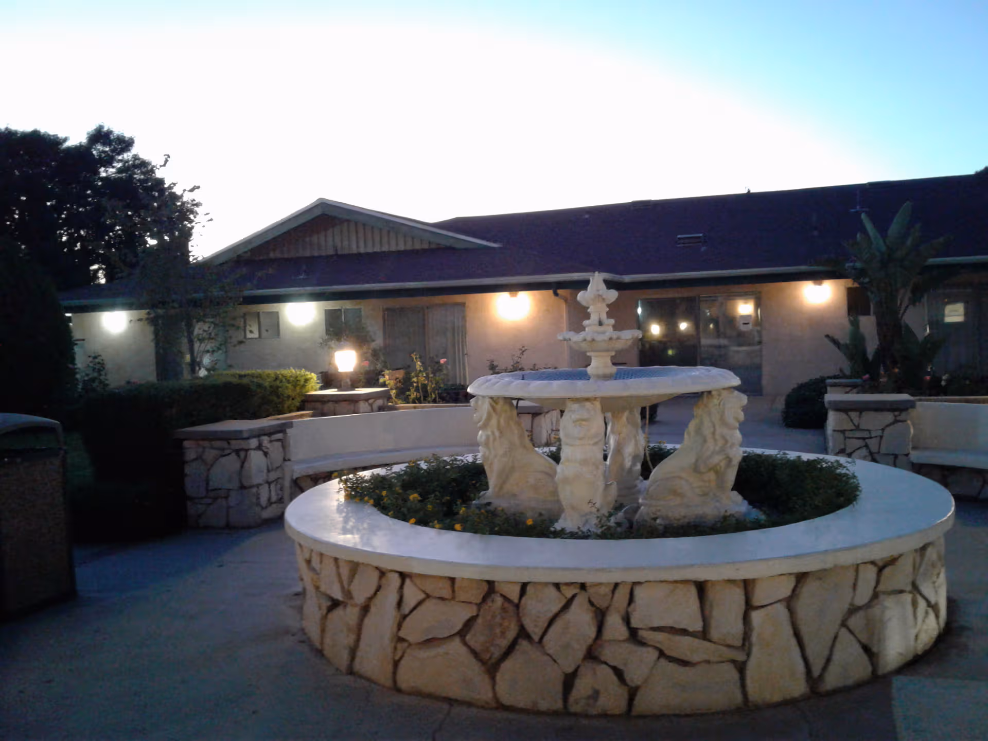 Outdoor courtyard featuring a stone fountain, seating walls, and a single-story facility in evening light.