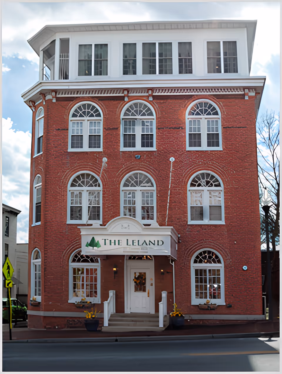 Front exterior of a red-brick, multi-story building with arched windows and a canopy sign reading 'The Leland' above the entrance.