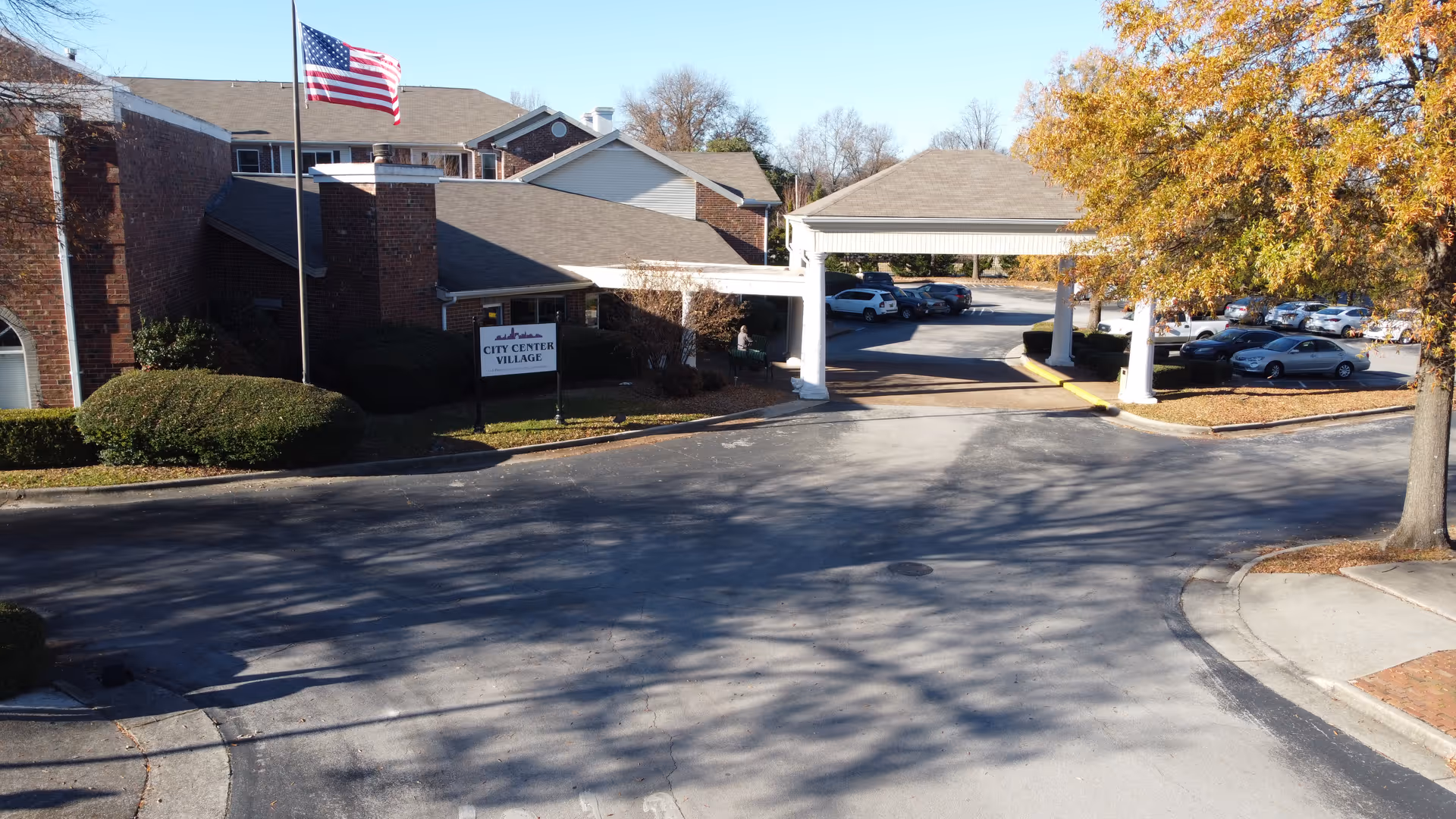 Exterior view of City Center Village Senior Community showing a brick building with an American flag on a flagpole, a covered entrance driveway, parked cars, and trees with autumn foliage.