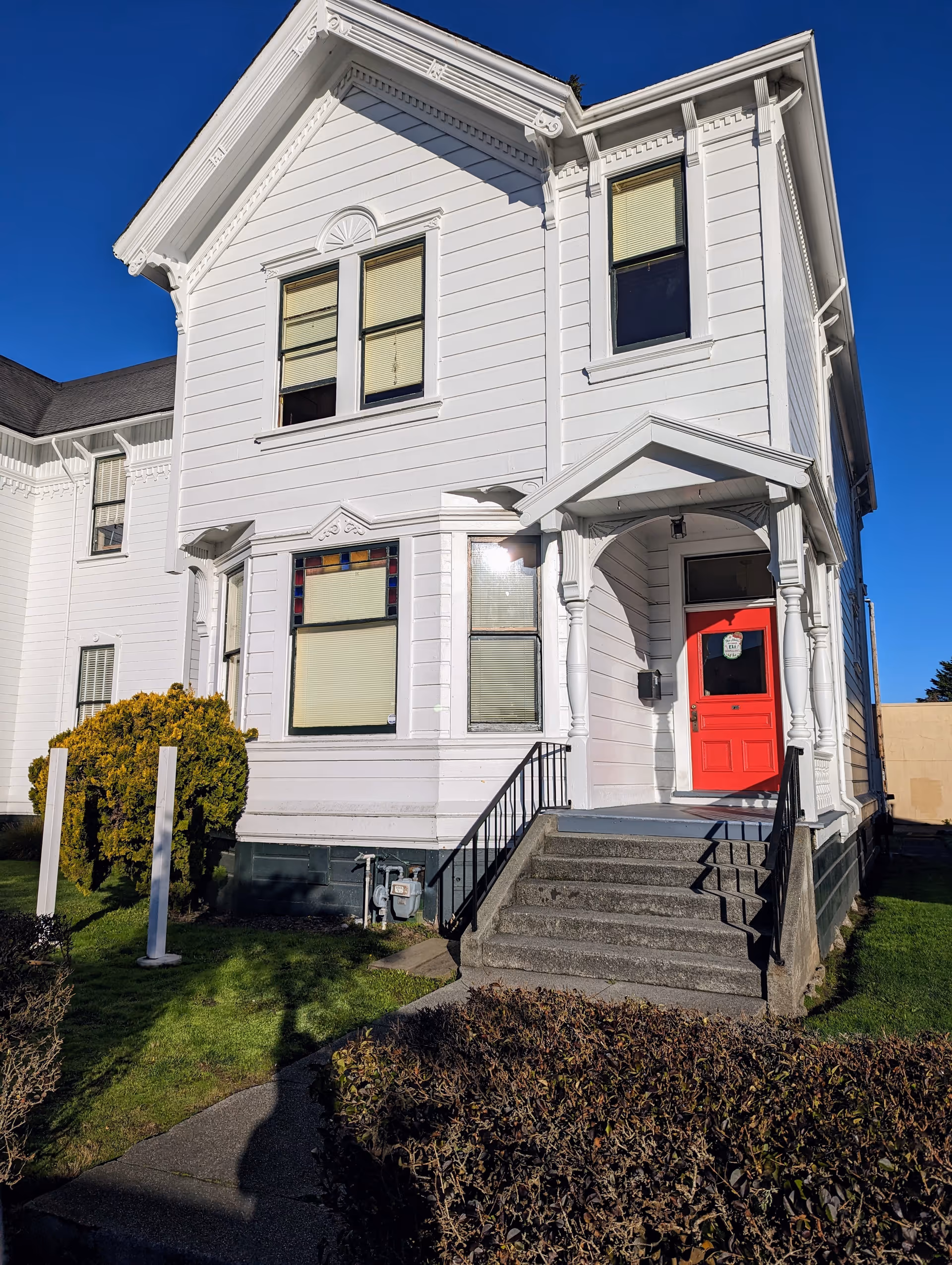 White wooden building with decorative trim, red front door, and concrete steps under a blue sky.