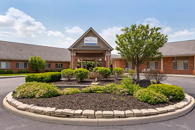 Front entrance of a brick senior living facility with a covered porte-cochere and a landscaped circular driveway island.