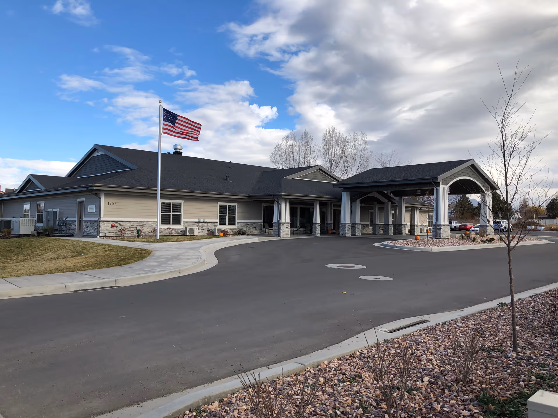 Exterior view of a single-story senior living facility building with a covered entrance driveway, an American flag on a flagpole, and a partly cloudy sky.