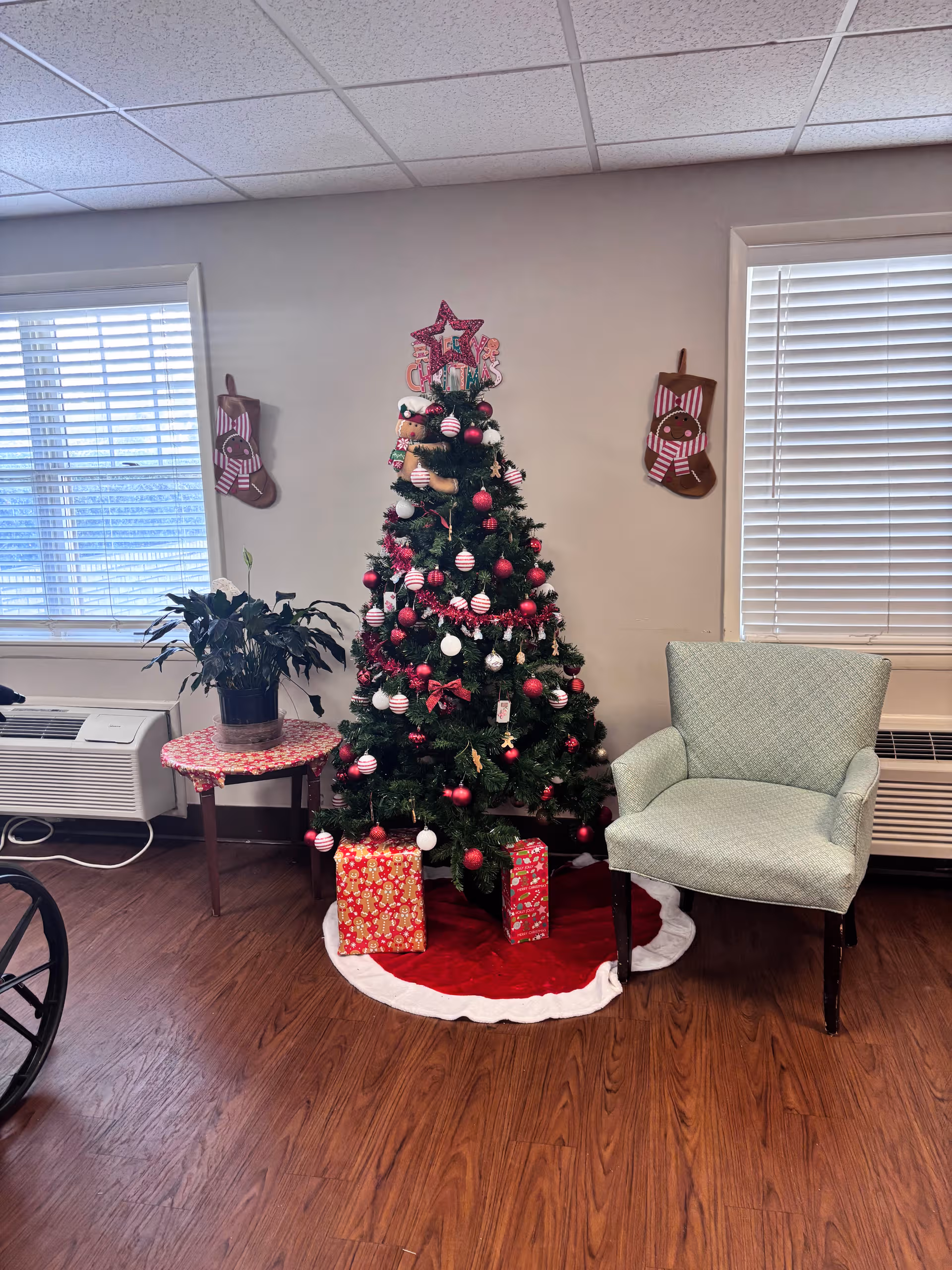 Seasonal Christmas tree with gifts beside a blue chair in a senior living room.