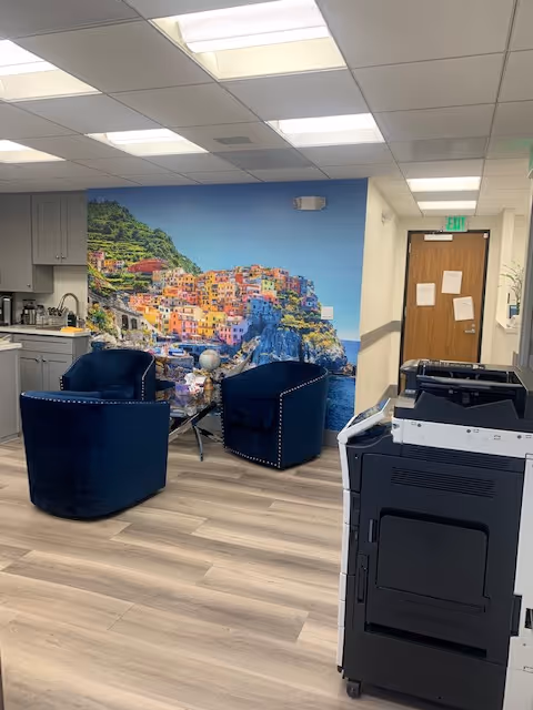 Interior lounge area with blue chairs, wall mural, copier, and kitchenette.