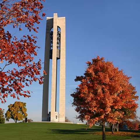 Tall bell tower on a grassy hill surrounded by autumn trees under a clear blue sky.