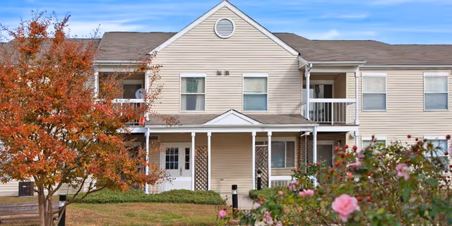 Exterior view of a beige two-story residential building with white trim, featuring a covered porch and balconies. There are flowering bushes and a tree with autumn-colored leaves in the foreground under a blue sky.