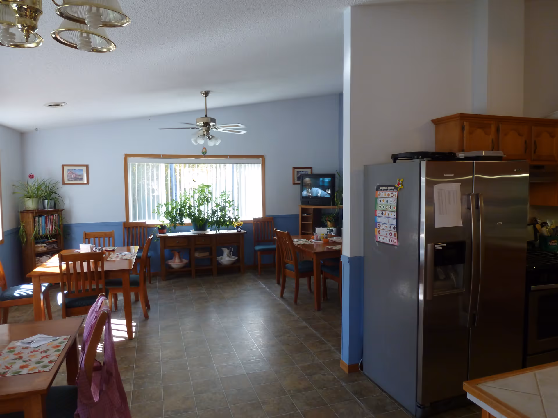 Senior living dining area with wooden tables, chairs, plants, and a stainless steel refrigerator.