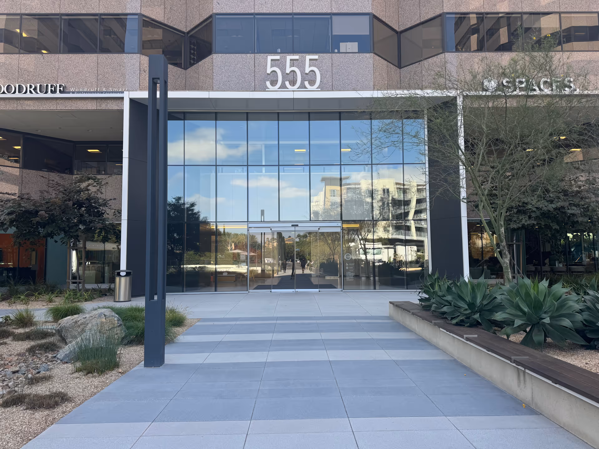 Glass-front entrance of a modern office building with surrounding desert landscaping and a walkway.
