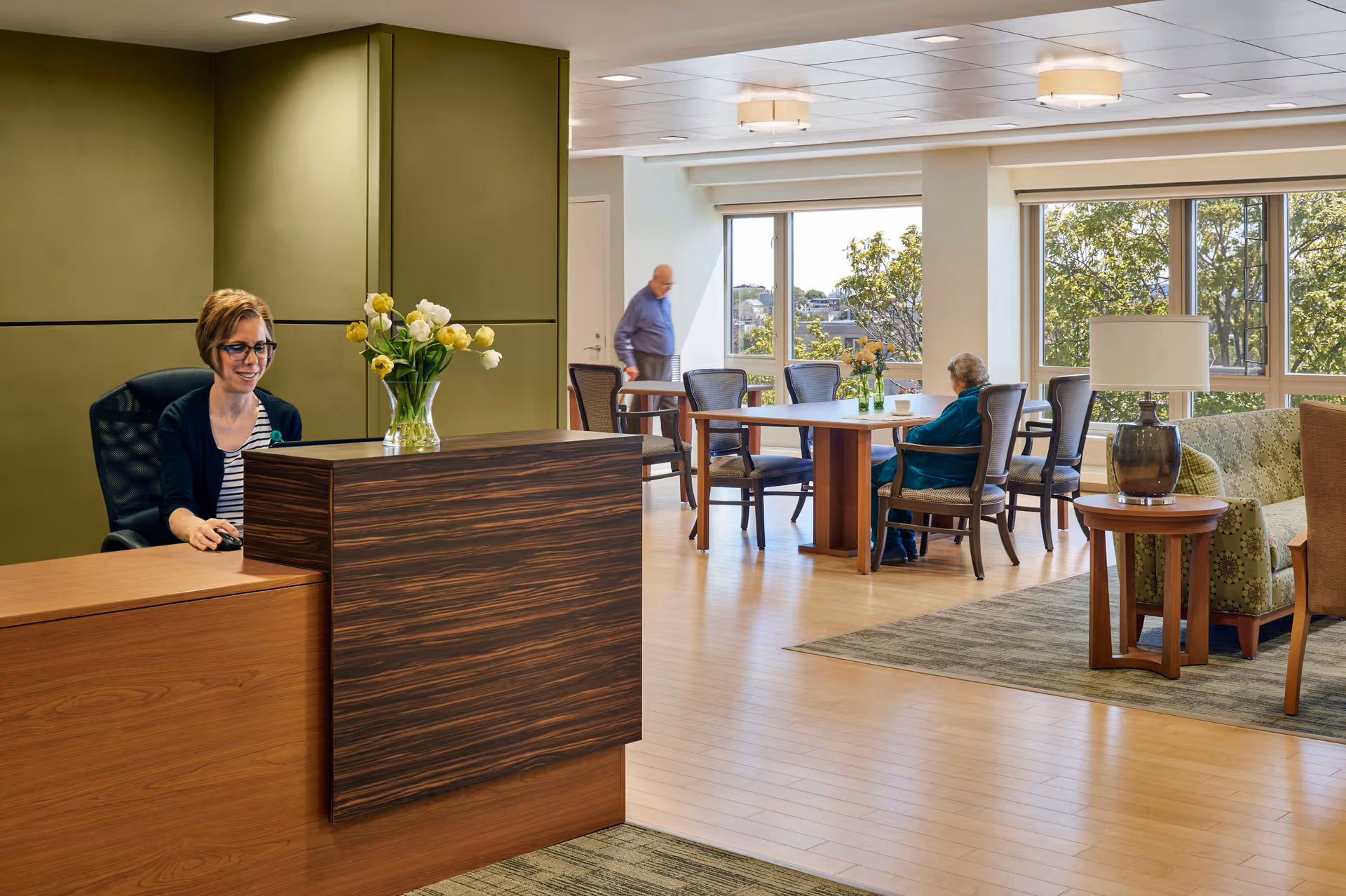 Receptionist sits at a wooden front desk in a bright senior living lobby.