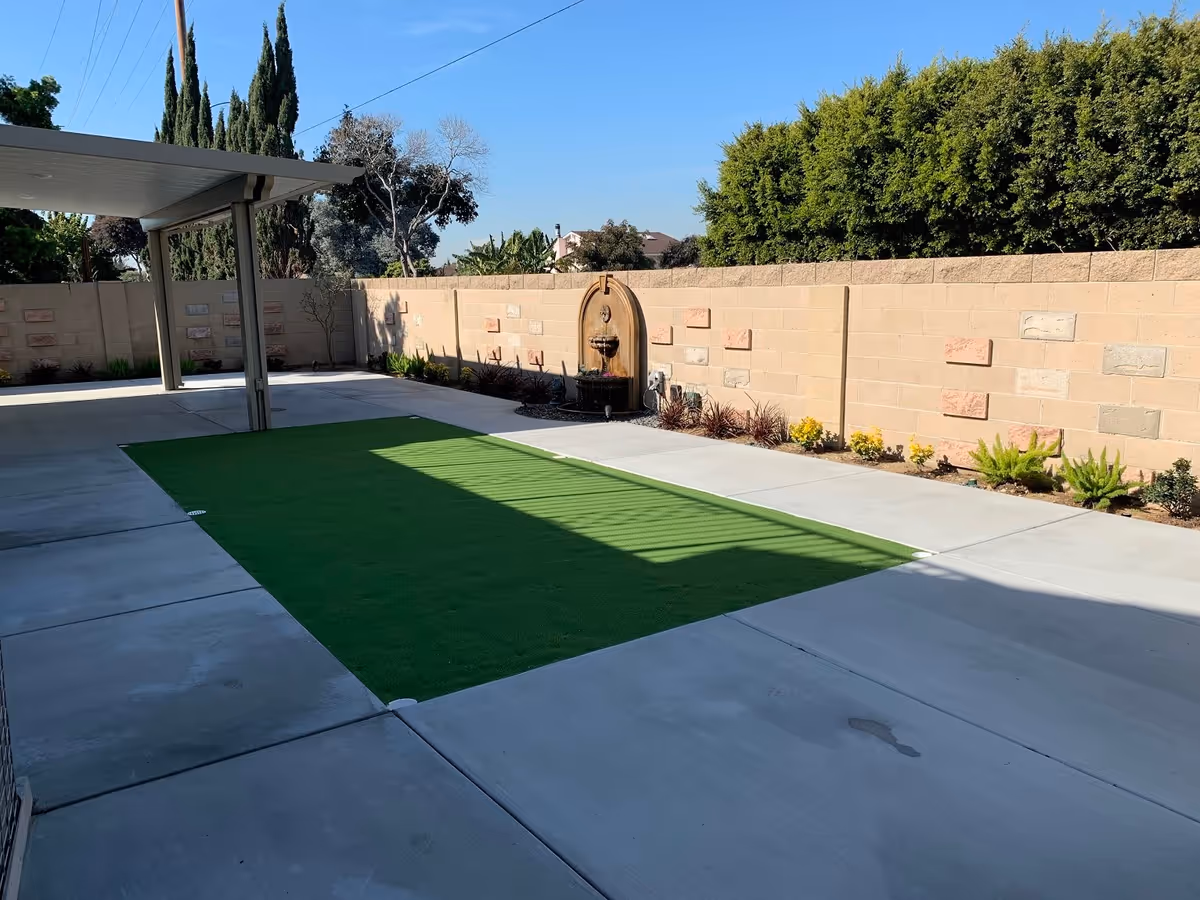Outdoor patio area with a covered section on the left, a rectangular patch of artificial grass in the center, and a decorative wall fountain mounted on a beige brick wall with small plants along the base of the wall under a clear blue sky.