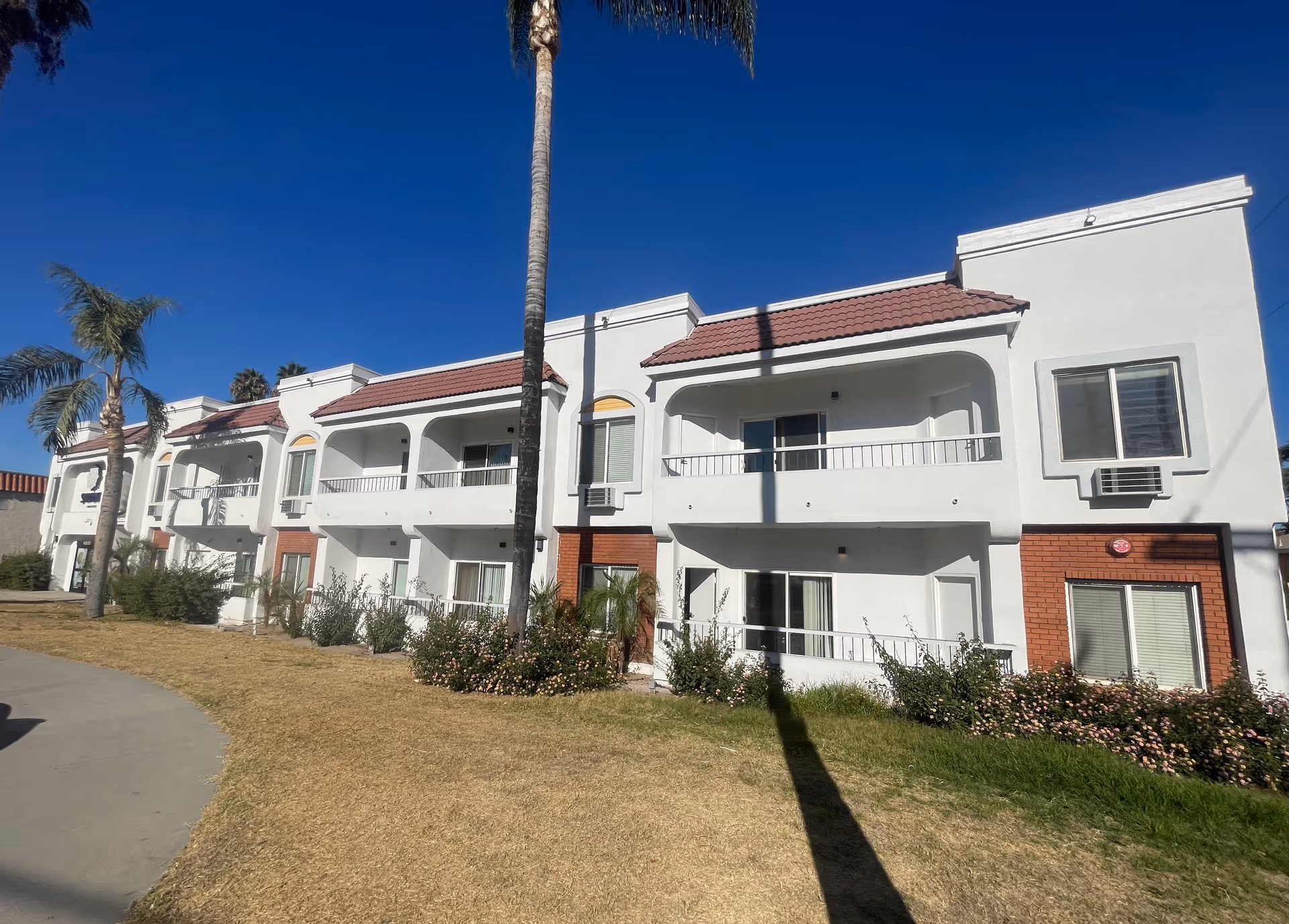 Two-story white residential building with balconies, red tile roof accents, palm trees and a grassy lawn under a clear blue sky.