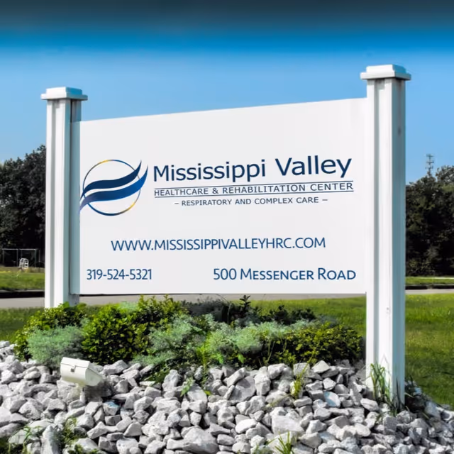 Large white roadside sign for Mississippi Valley Healthcare & Rehabilitation Center with logo, website, phone number, and address surrounded by landscaping and rocks.