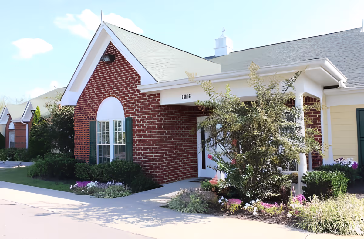 Exterior view of a senior living facility building with red brick walls, white trim, green window shutters, and a small garden with bushes and flowers near the entrance under a clear blue sky.