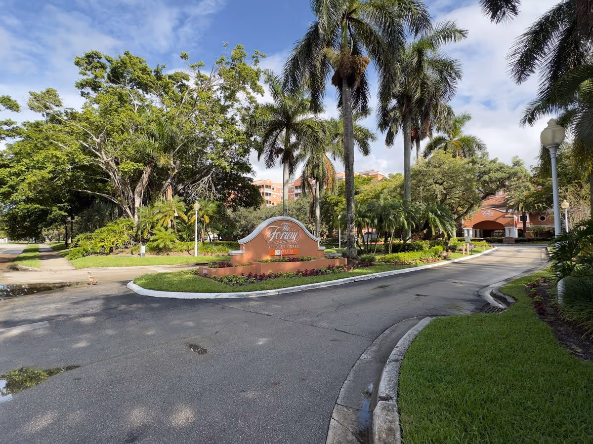 Entrance drive and landscaped sign for The Forum at Deer Creek with palm trees and the facility building visible behind.