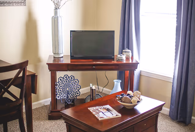 A cozy living room corner featuring a wooden TV stand with a flat-screen TV, decorative vases, and a patterned ornament. In front, a wooden coffee table holds a bowl with decorative balls and a magazine. The room has beige walls, carpeted floor, and a window with blue curtains letting in natural light.