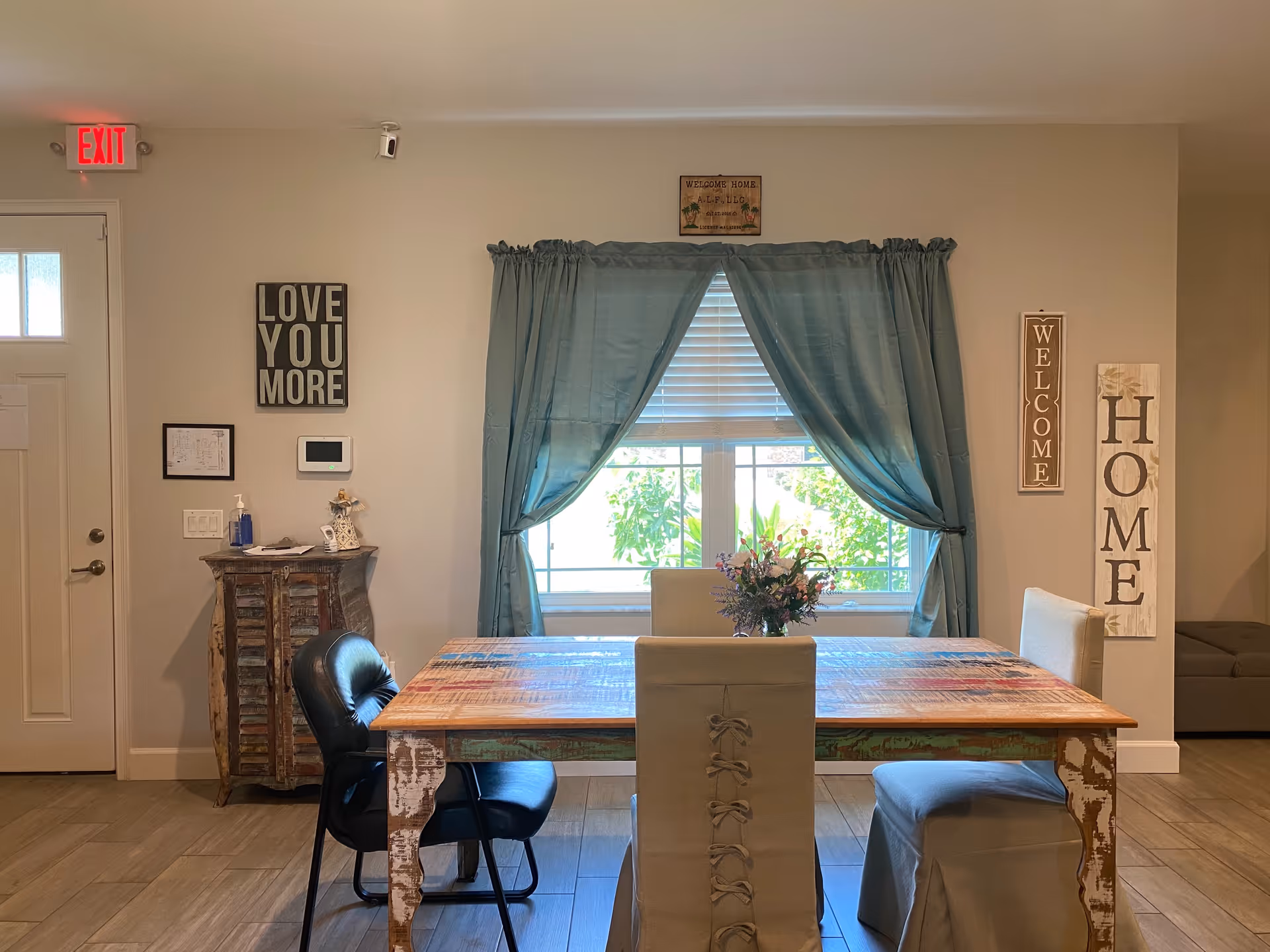A cozy dining area with a rustic wooden table surrounded by four chairs, two with white covers and two black leather chairs. Behind the table is a window with blue curtains tied back, letting in natural light and showing greenery outside. On the walls are decorative signs including 'LOVE YOU MORE', 'WELCOME', and 'HOME'. A small wooden cabinet with hand sanitizer and papers sits near a white door with an exit sign above it.