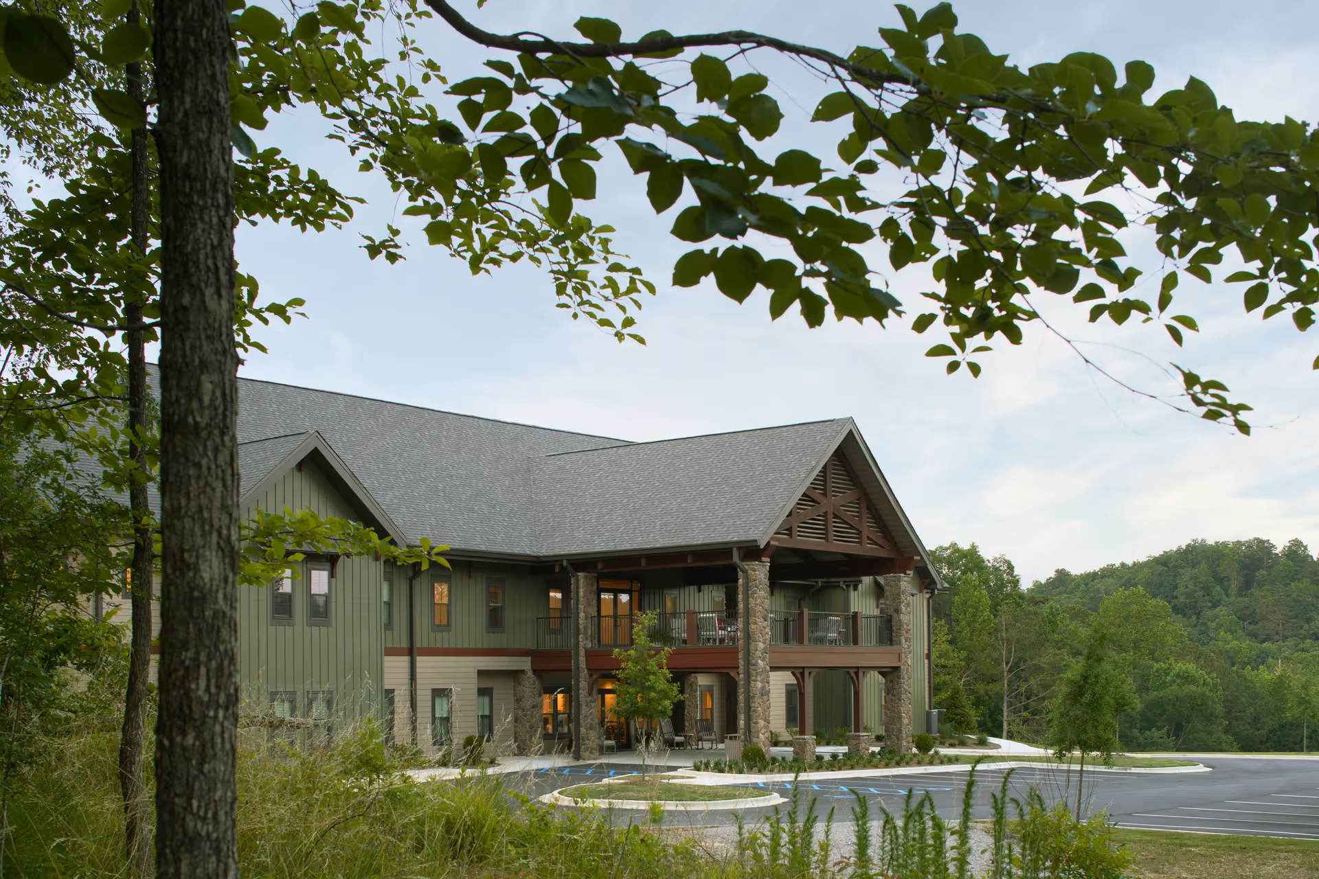 Exterior view of a two-story building with a large covered porch supported by stone columns, surrounded by trees and greenery under a partly cloudy sky.