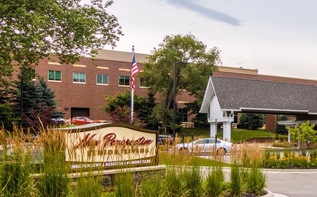 Exterior view of New Perspective Senior Living | Woodbury facility with a brick building, an American flag on a flagpole, a covered entrance, and landscaping with tall grasses and trees.
