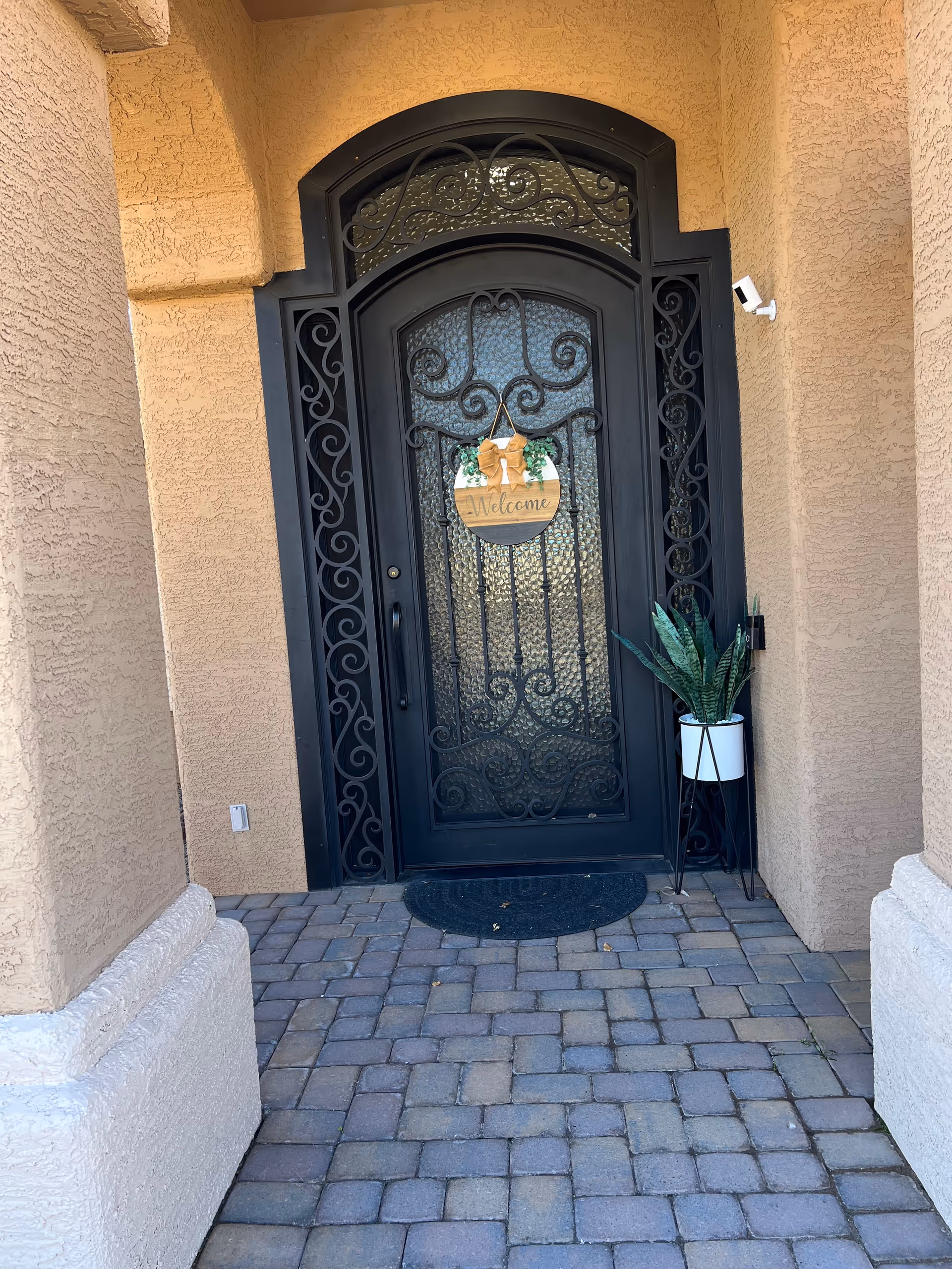 Arched wrought-iron front door with a welcome sign, potted plant, and beige exterior.