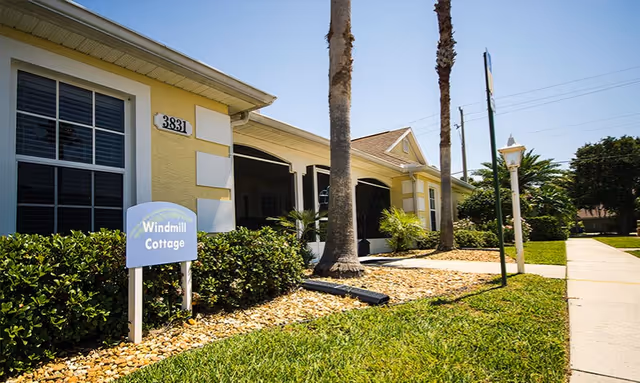 Front exterior of a yellow single-story cottage with a "Windmill Cottage" sign, palm trees, and a sidewalk.