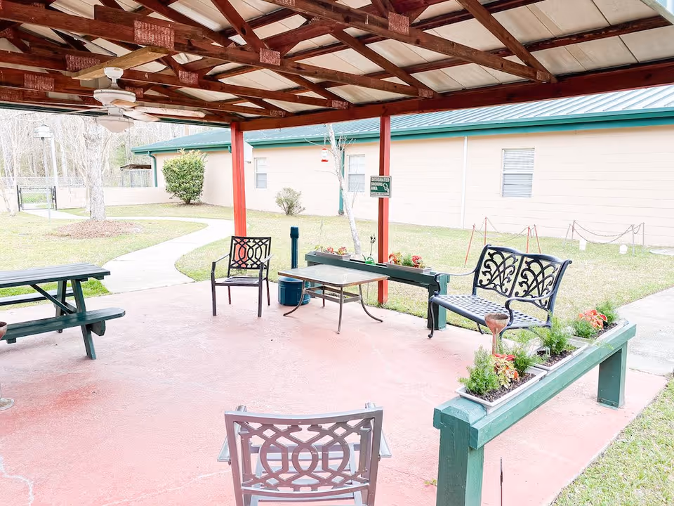 Covered outdoor patio area with metal chairs, a metal bench, a glass-top table, and a green picnic table. The patio has a wooden roof with ceiling fans and overlooks a grassy yard with a beige building in the background.
