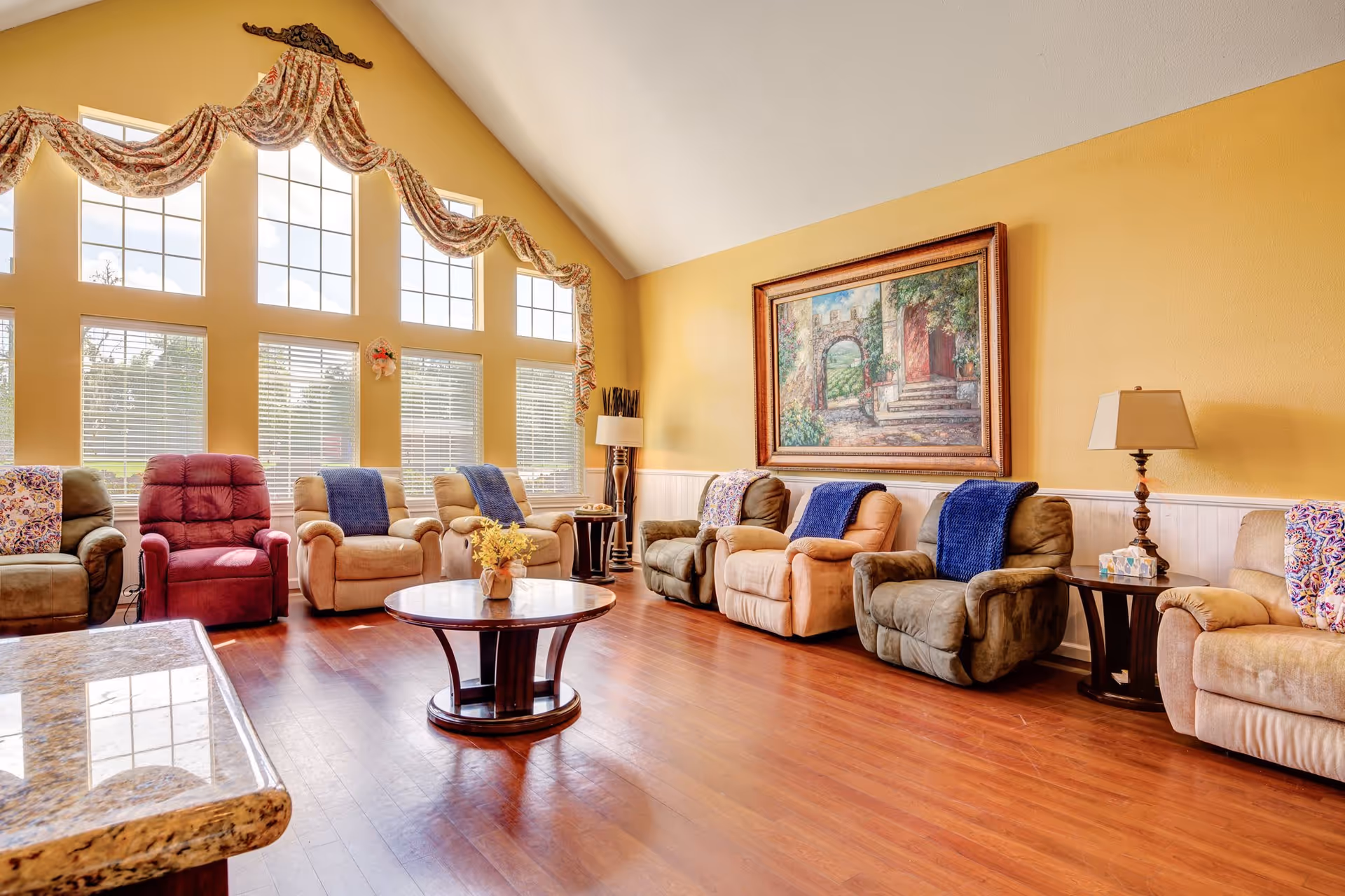 Sunlit communal living room with a row of recliner chairs, large tall windows, a central coffee table, and framed artwork on a yellow wall.