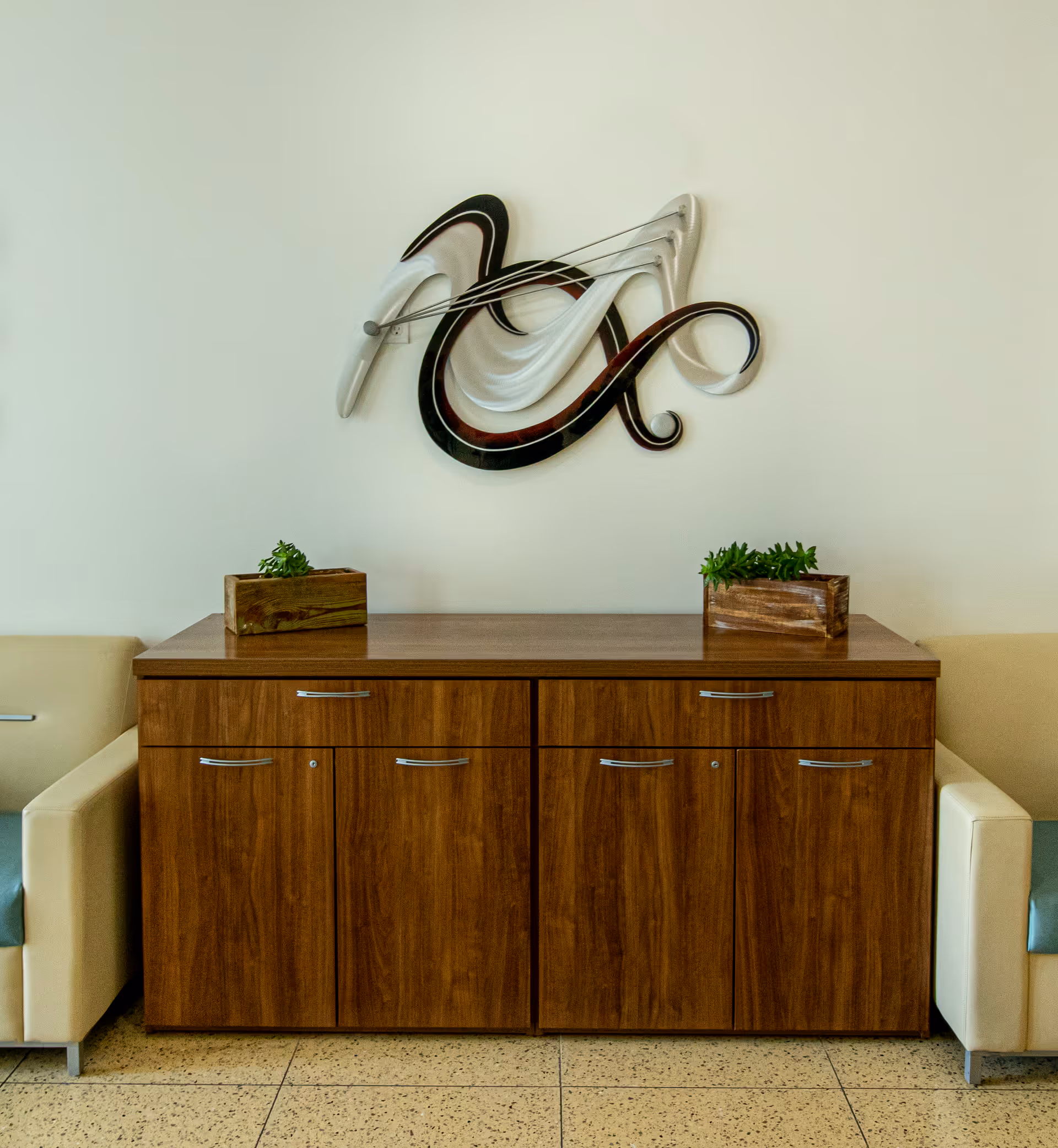 A wooden cabinet with six drawers and doors, flanked by two cream-colored armchairs with teal cushions. On top of the cabinet are two small wooden planters with green plants. Above the cabinet is a modern abstract wall sculpture with flowing curves in black, white, and brown tones mounted on a plain light-colored wall.