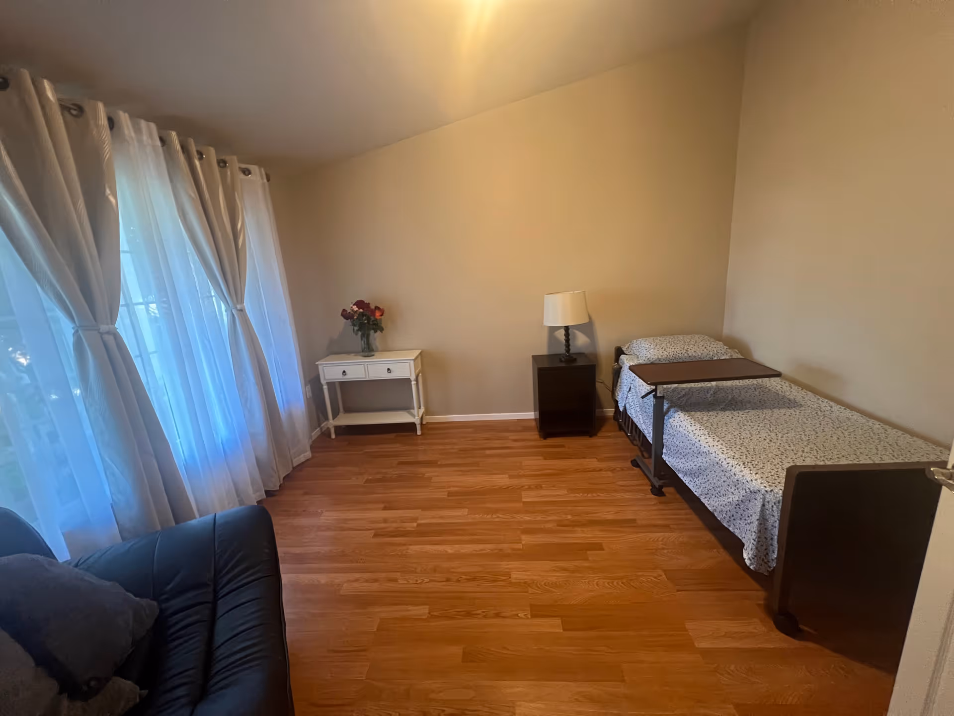 A small bedroom with a single bed, nightstand, lamp, and sheer white curtains by a window.