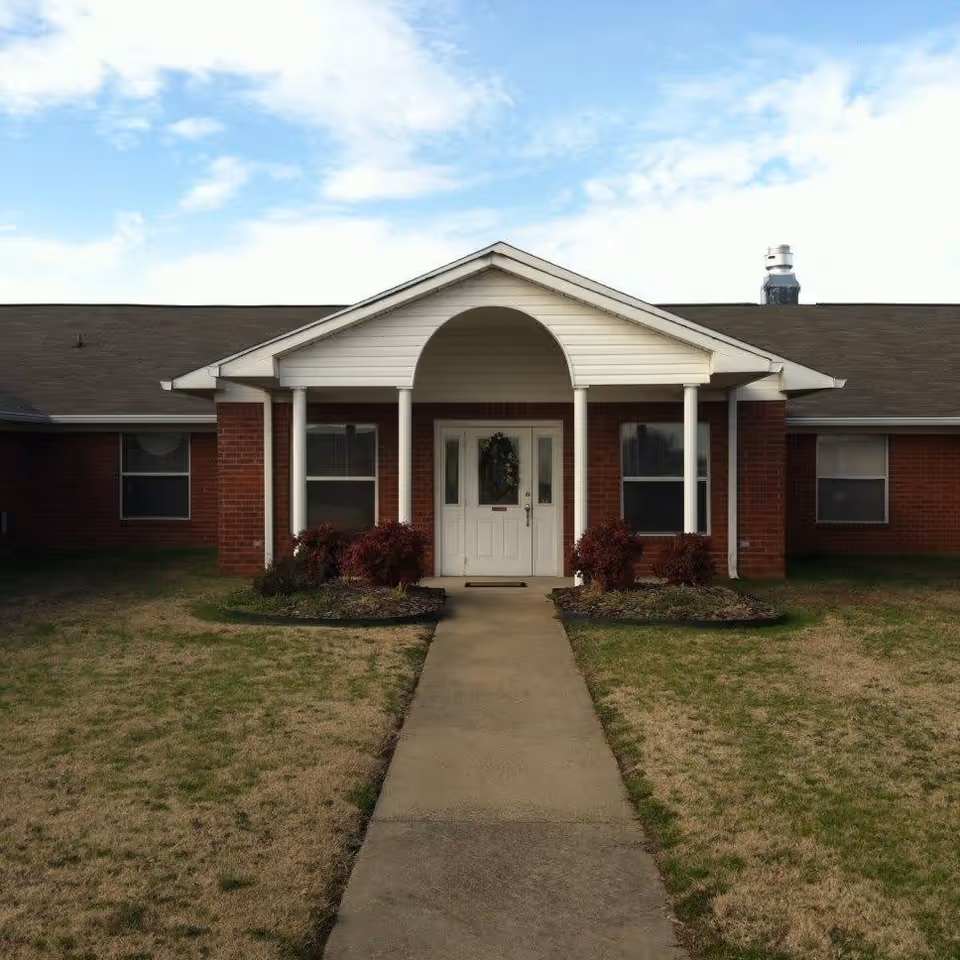 Front exterior view of a single-story brick building with a white entrance door under a small porch supported by four white columns. There are two windows on either side of the door and small landscaped areas with bushes along the walkway leading to the entrance. The sky is partly cloudy.