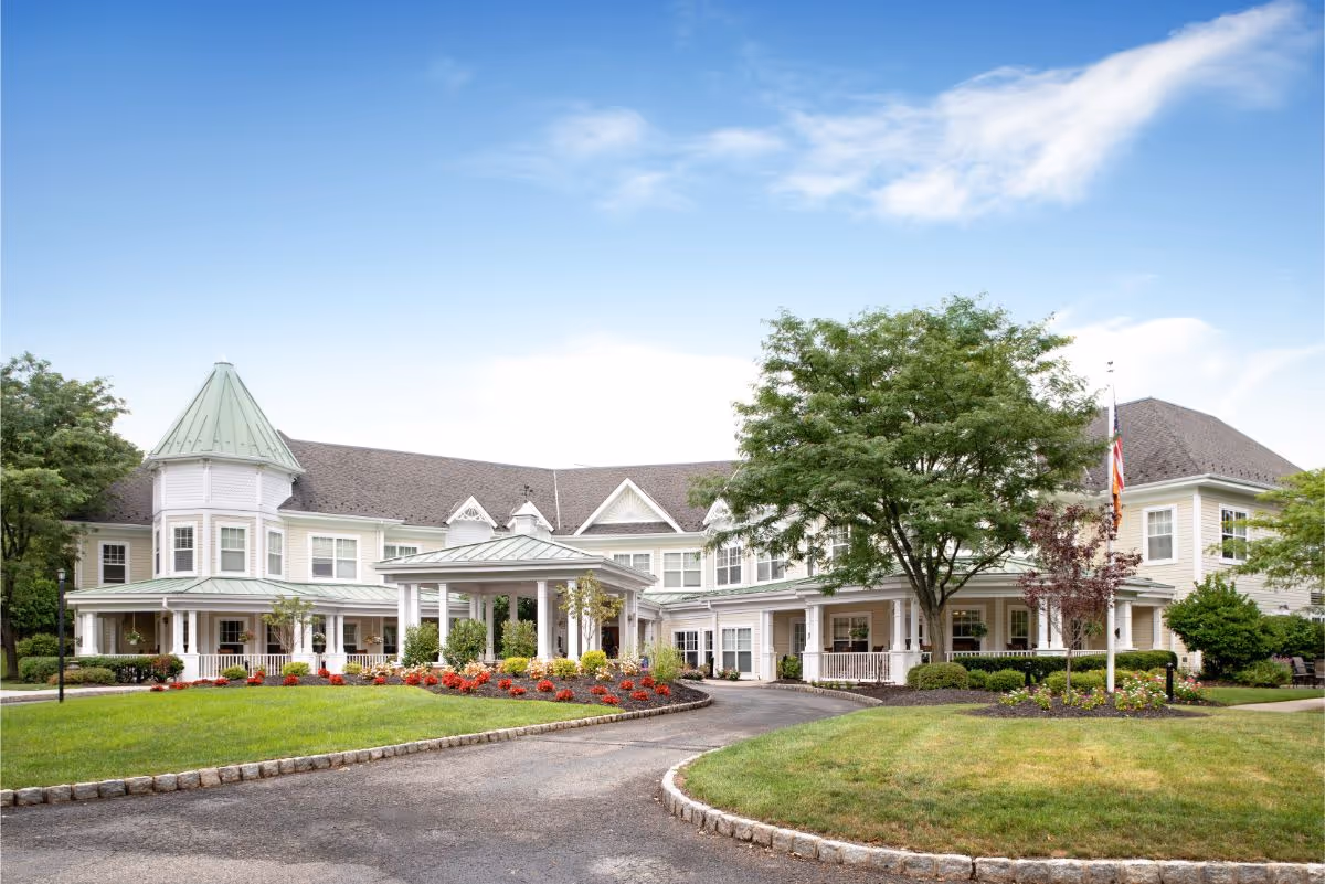Exterior view of a large, two-story senior living facility building with a green roof, white siding, and a covered entrance. The building is surrounded by well-maintained landscaping including green lawns, trees, and flower beds under a partly cloudy sky.