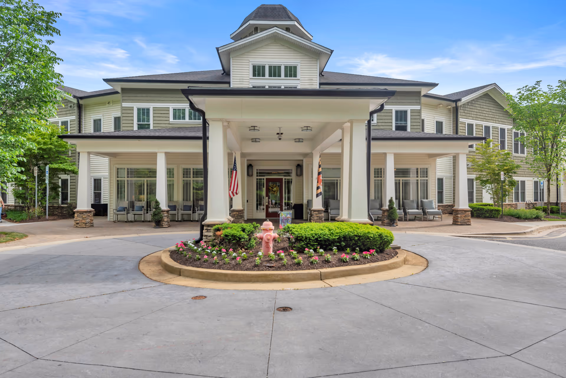 Front exterior view of a senior living facility with a covered entrance, white columns, and a circular driveway. The building is two stories with beige siding and multiple windows. There are American and Maryland state flags near the entrance, and landscaped greenery with flowers and a pink fire hydrant in the center of the driveway circle.