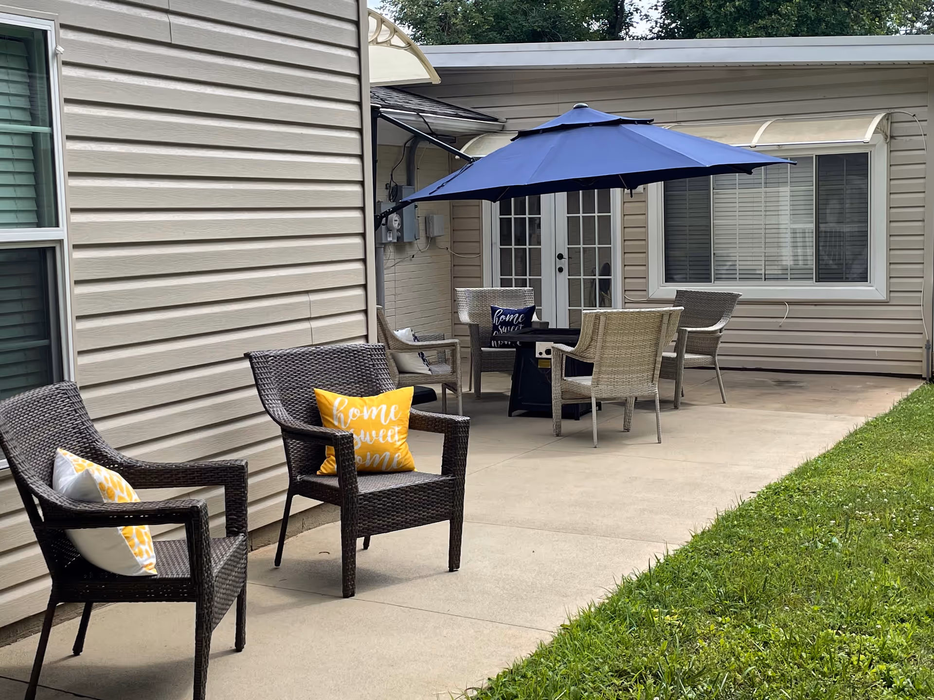 Outdoor patio with wicker seating and a blue umbrella beside a beige senior living home.
