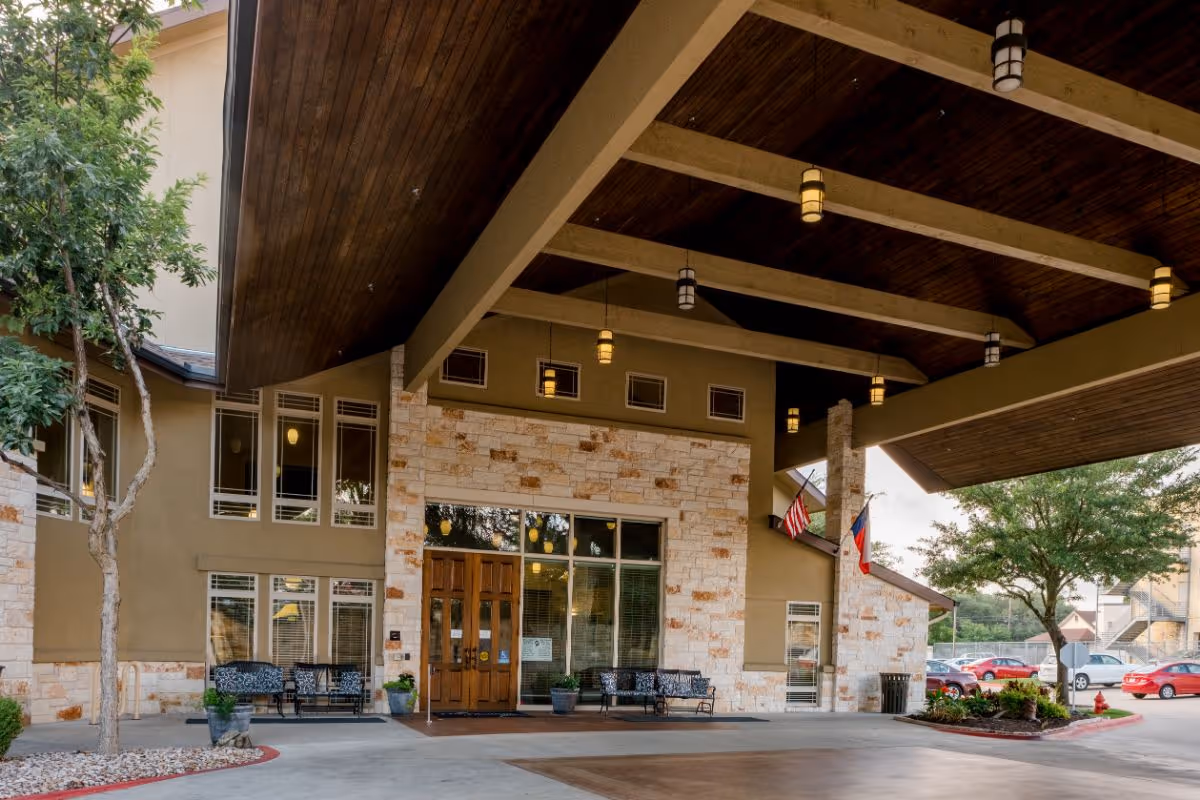 Covered main entrance of a senior living facility featuring a large wooden canopy, stone facade, glass doors, benches, hanging lights, and flags.