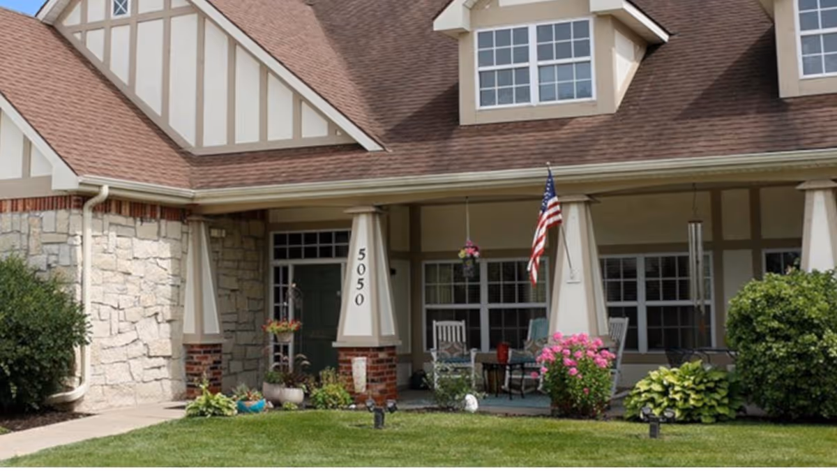 Front exterior view of a building with a porch featuring white pillars, an American flag, hanging flower pots, rocking chairs, and well-maintained landscaping with green grass and bushes.