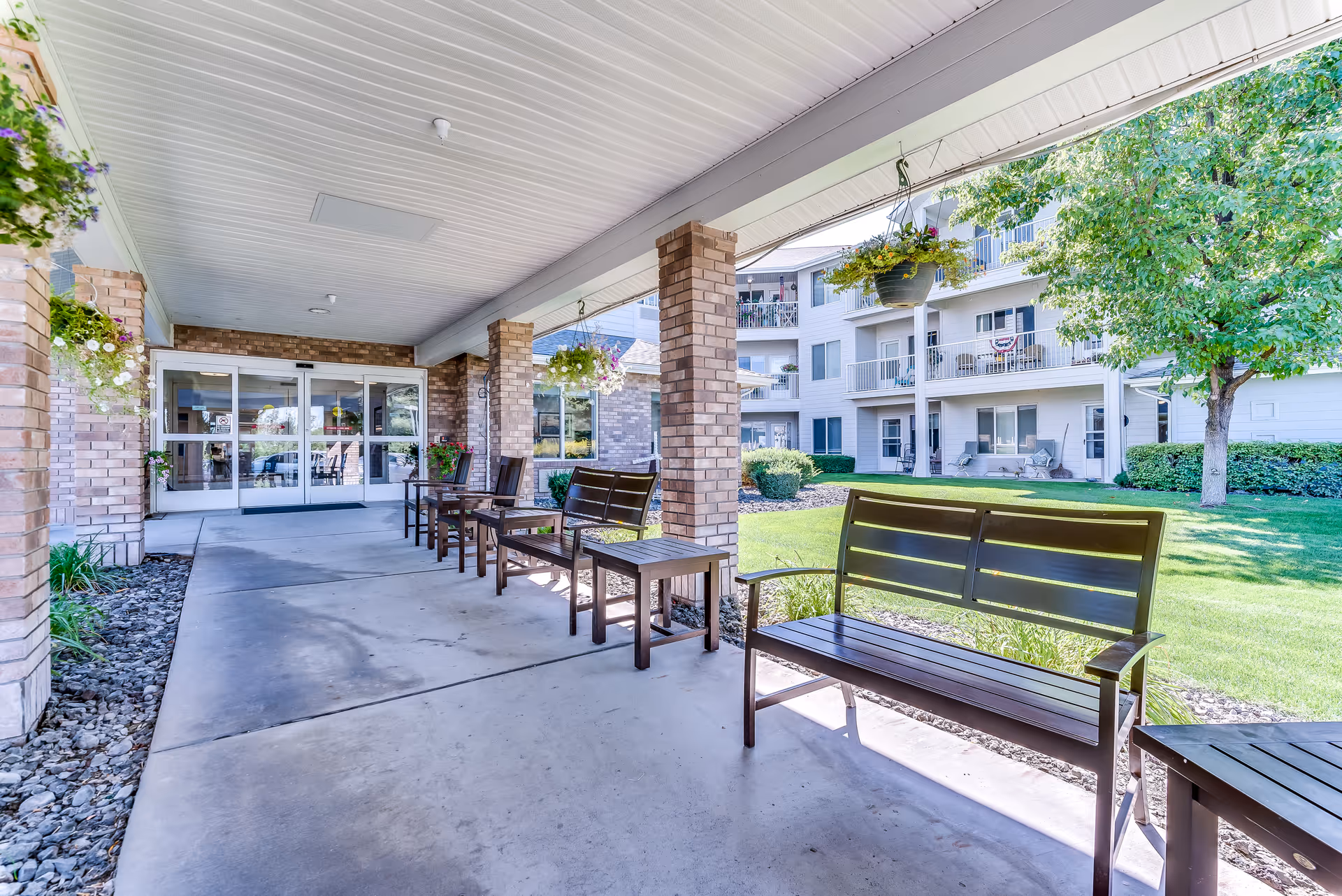 Covered front entrance with benches along a walkway facing the lawn and multi-story senior living building.