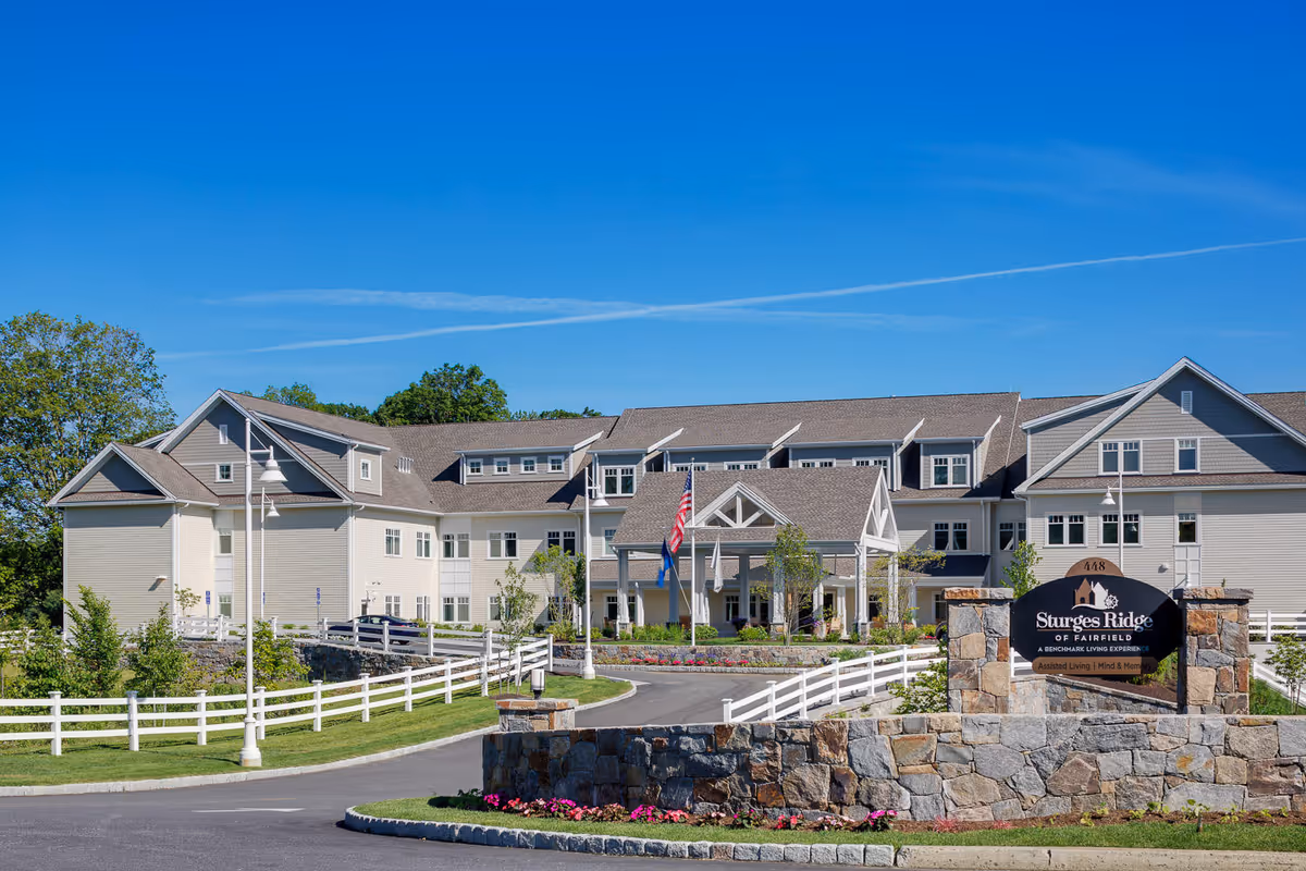 Exterior view of Sturges Ridge of Fairfield, a large assisted living and memory care facility with beige siding and multiple windows. The building has a covered entrance with white pillars and an American flag flying in front. There is a stone sign in the foreground with the facility's name and description. The area is landscaped with green grass, trees, and flowers under a clear blue sky.