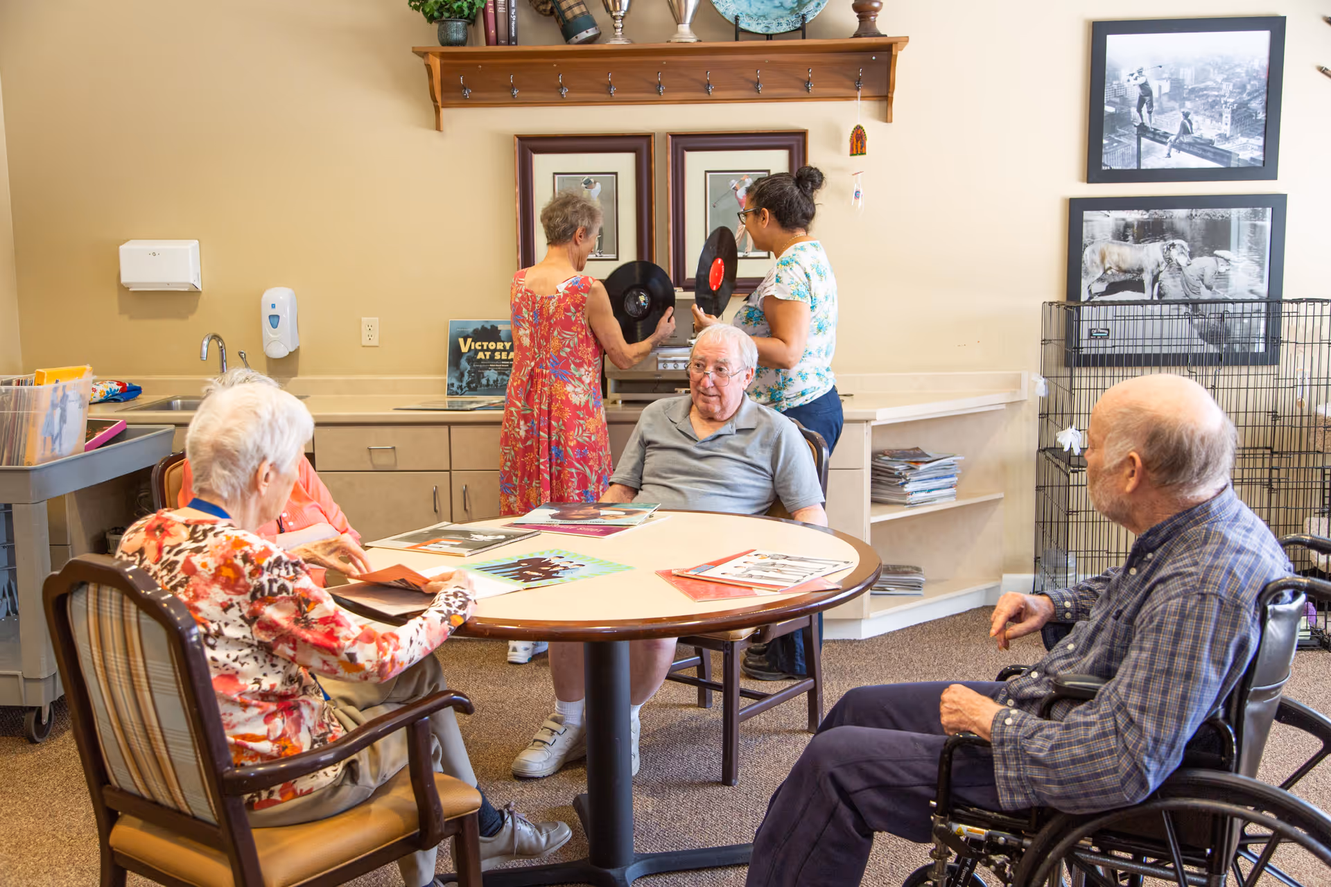 A group of elderly people in a communal room at an assisted living facility. Three seniors are seated around a round table looking at records and papers, while two women stand near a counter holding vinyl records. The room has beige walls, framed pictures, and a shelf with books and decorative items.