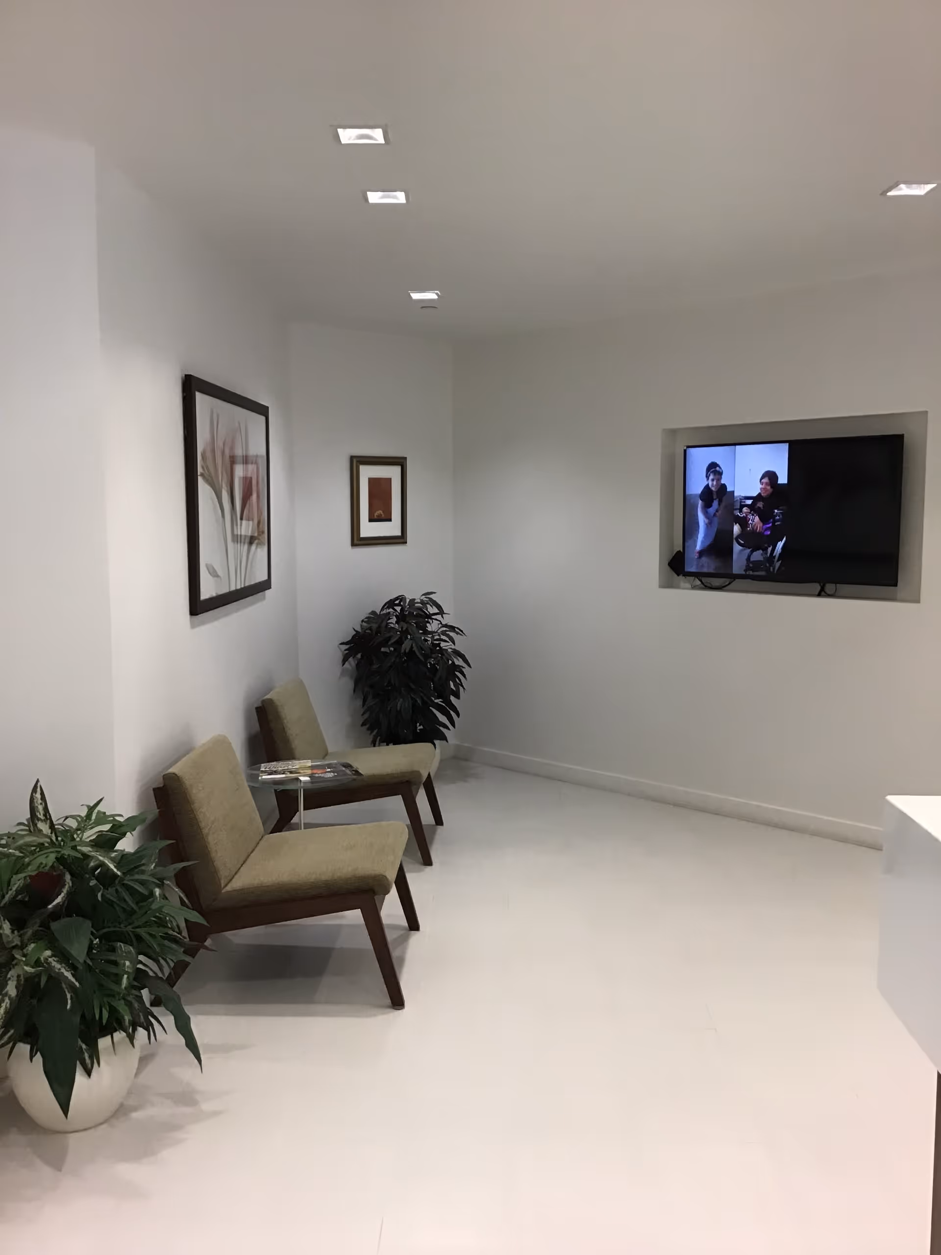 Waiting area with two beige chairs, magazines, potted plants, wall art, and a mounted TV.