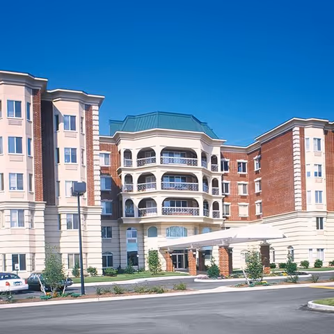 Exterior view of a multi-story assisted living facility with a covered entrance, balconies, and a mix of brick and light-colored siding under a clear blue sky.