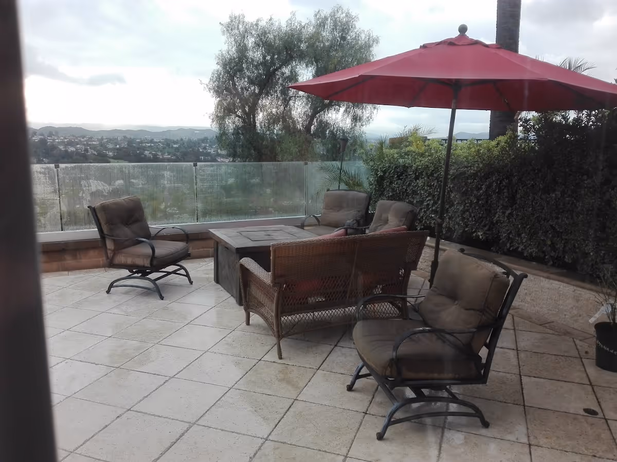 Outdoor patio with cushioned chairs, a wicker loveseat, a red umbrella, and a glass railing overlooking a distant landscape.