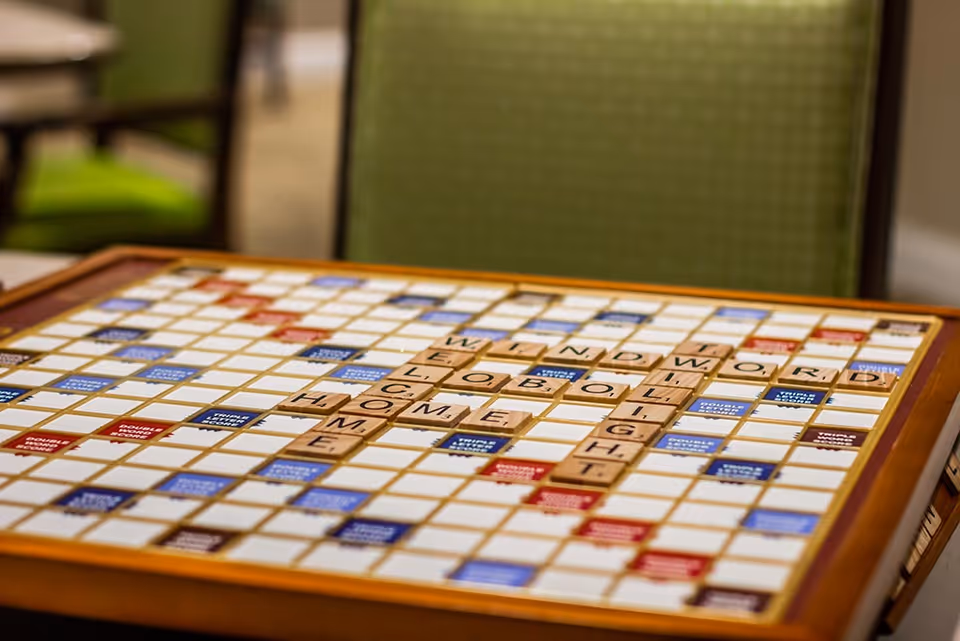 Close-up of a Scrabble board with wooden letter tiles spelling words like WINDOW, WORLD, LIGHT, HOME, and LOB. The board is on a table with a green cushioned chair in the background.