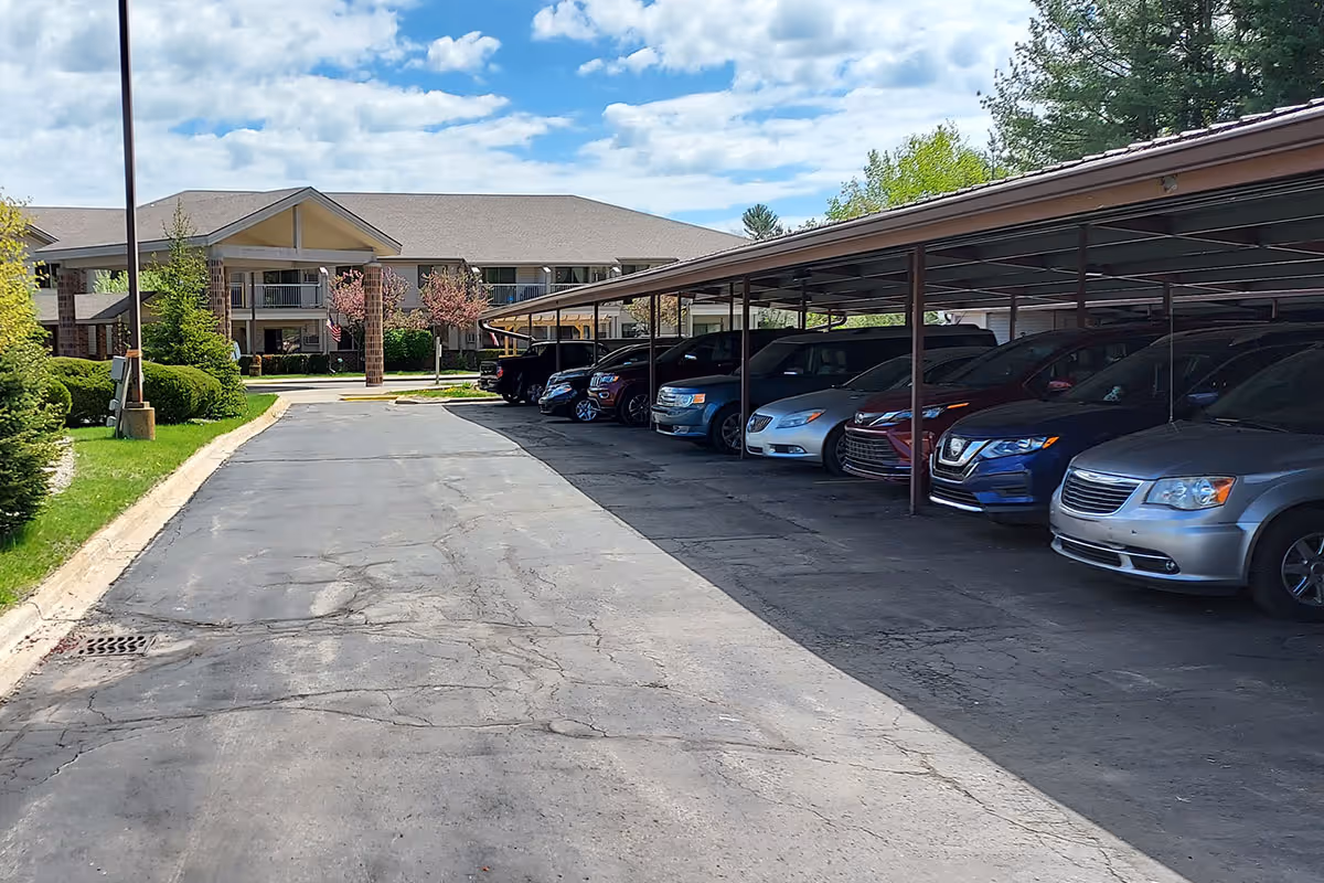 View of a senior living facility parking area with a covered carport on the right side and a two-story building with balconies in the background under a partly cloudy sky.