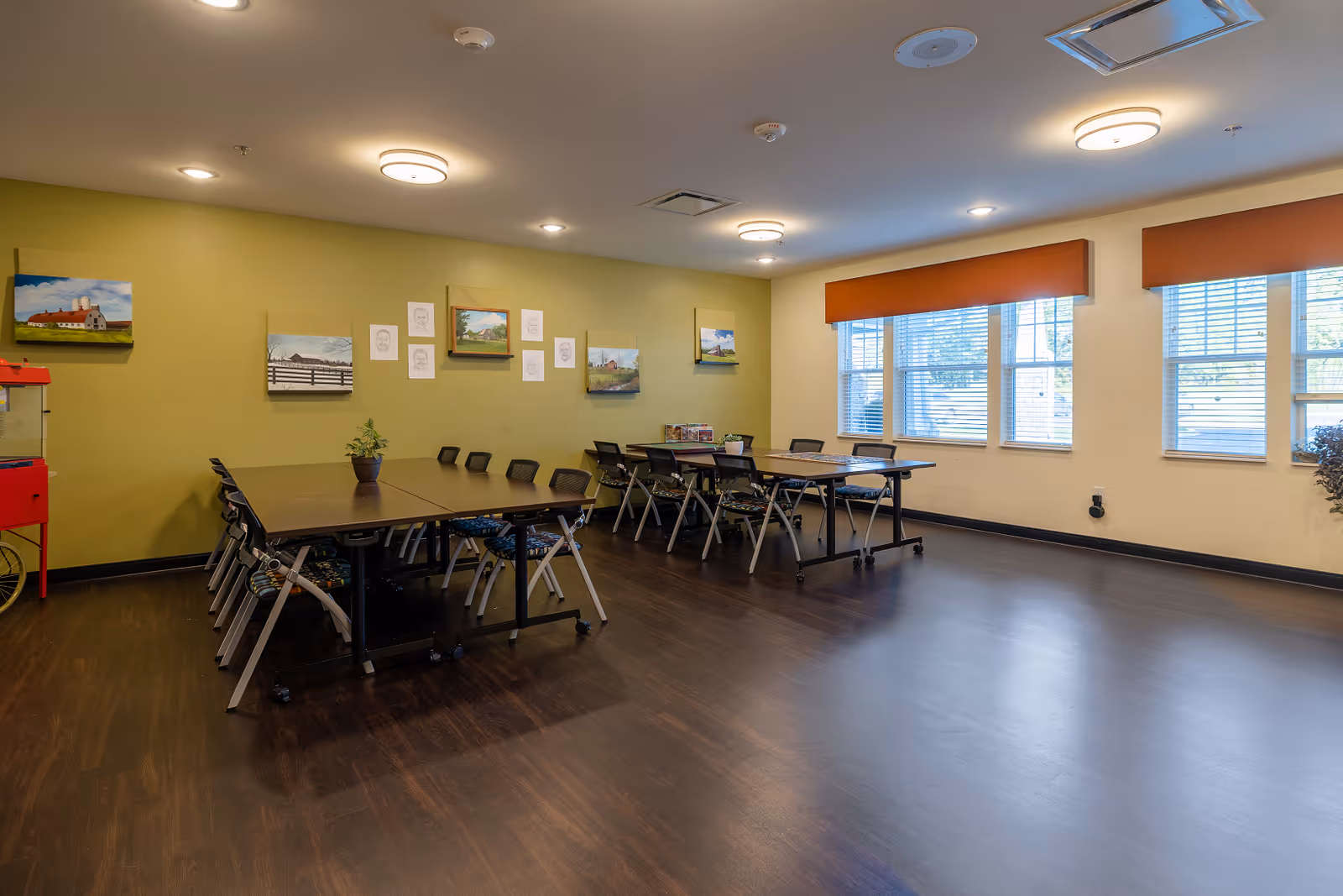 Bright multipurpose dining area with long tables, chairs, and wall art in a senior living facility.