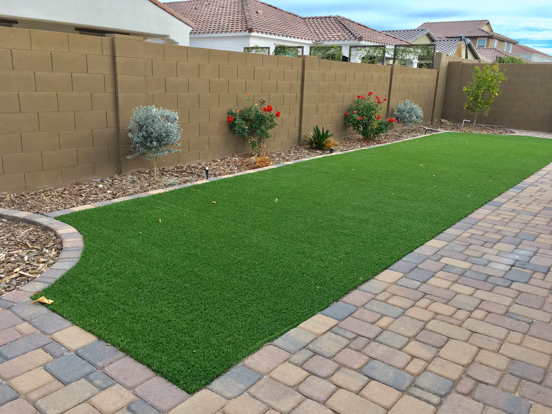 A well-kept backyard lawn with a paver walkway and shrubs along a block wall.