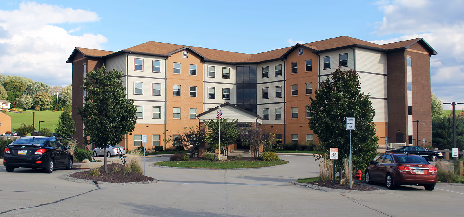 Four-story brick-and-siding care facility with a covered entrance, circular driveway and parked cars under a partly cloudy sky.