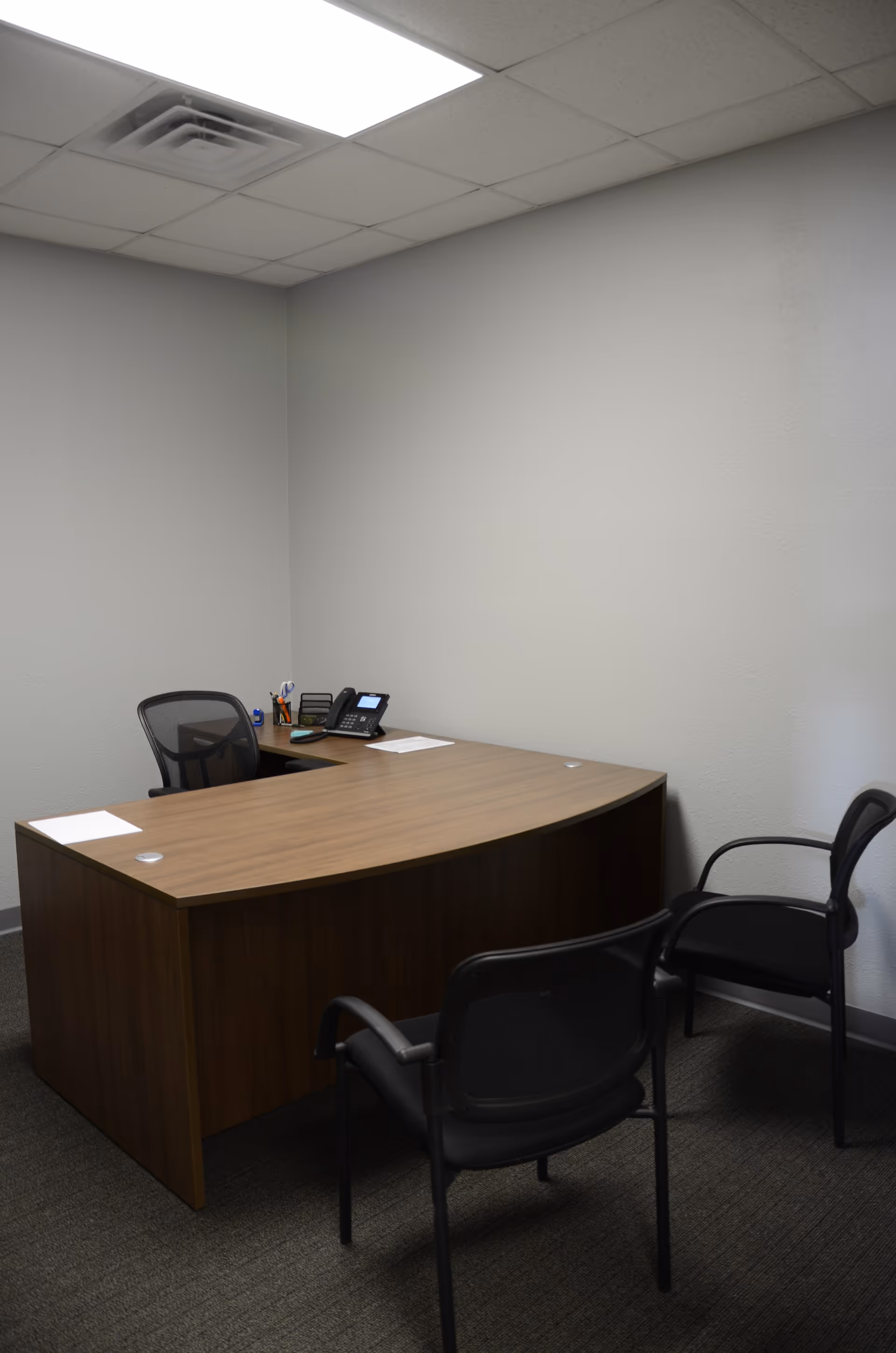A small office with a wooden curved desk, chairs, and office supplies against gray walls.