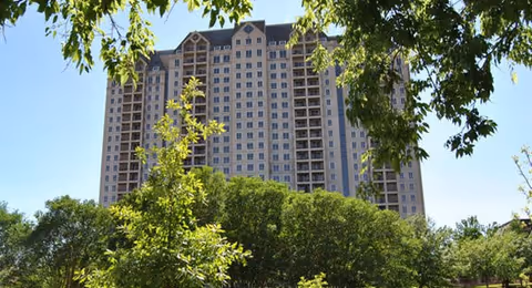 Tall beige residential tower framed by green trees and blue sky.