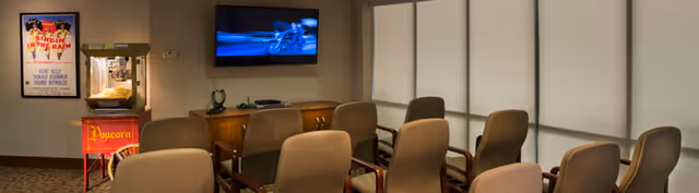 A small theater room with several rows of beige chairs facing a wall-mounted flat screen TV. To the left, there is a popcorn machine and a framed Singin' in the Rain movie poster on the wall. The room has large windows covered with blinds on the right side.