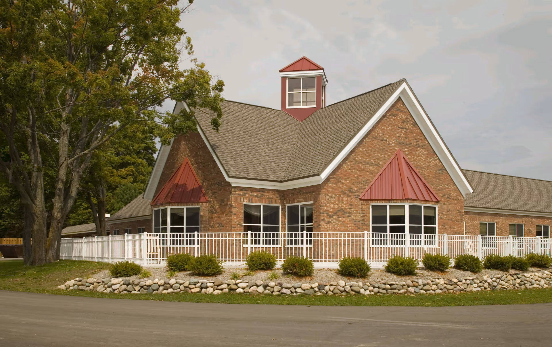 Brick senior living facility exterior with white railing, shrubs, and a stone border.
