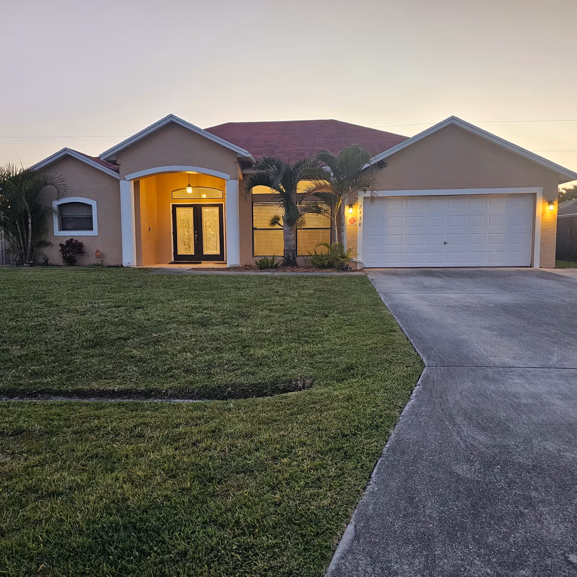 A single-story beige house with a two-car garage and manicured lawn at dusk.